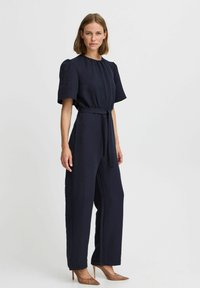 Tuta blu navy con tessuto testurizzato, maniche corte a palloncino, vita aderente accentuata da una cintura e gambe larghe. Completato da tacchi beige.