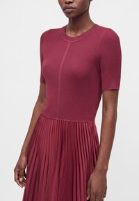 Femme portant un haut côtelé à manches courtes avec une robe jupe plissée d’un rouge profond, montrée de l’épaule jusqu’à mi-cuisse sur un fond uni.