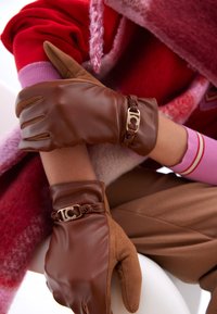 Bruna läderhandskar med flätad detalj och logga. Handskarna har en tät passform och avsmalnande fingrar, kombinerade med ett strukturerat röd- och rosalinnande klädesplagg.