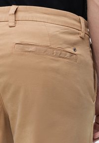 Personne portant un pantalon beige vue de dos, avec un gros plan sur une poche arrière passepoilée et un détail de logo métallique au-dessus de la poche.