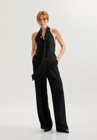 Haut noir à col halter avec un nœud à l'avant et une petite épingle décorative, associé à un pantalon noir ample et des escarpins noirs à bout pointu.