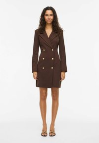 Ženska stoji, oblečena v rjavo obleko-blazer z dvojnim zapenjanjem in zlatimi gumbi ter črnimi čevlji s odprtimi prsti, na enostavni belem ozadju.