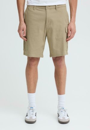 Mężczyzna ubrany w beżowe krótkie spodnie bojówki, białe skarpetki za kostkę i białe trampki z czarnymi paskami, stojący na jednolitym tle.