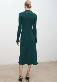 Vestido de manga larga, color verde bosque, con corsé ajustado, dobladillo fluido y un sutil detalle de cremallera en la parte trasera; combinado con sandalias de tacón negras.