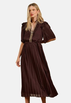 Blonde Frau trägt ein dunkelbraunes plissiertes Midikleid mit Rüschen- und verzierten Details am V-Ausschnitt und an den kurzen Ärmeln.