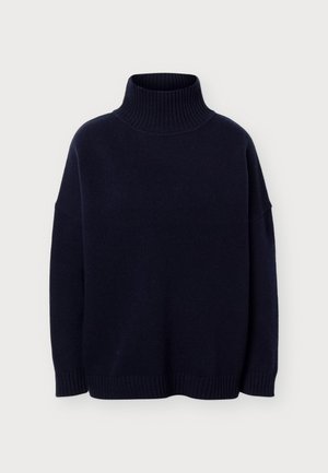 Persona de pie con un jersey de cuello alto negro, pantalones beis anchos y zapatos blancos sobre un fondo claro y liso.