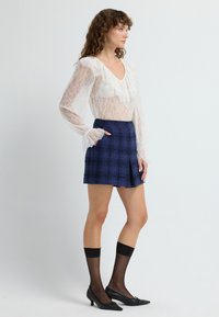 Blusa di pizzo con maniche lunghe e scollo arricciato; gonna a quadri blu con pince; décolleté neri; calze nere trasparenti.