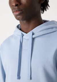 Sudadera azul clara hecha de un tejido suave. Presenta una capucha con cordón, acentos texturizados en las tiras y el logo "HUGO" en una impresión sutil.
