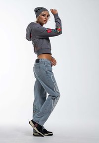 Sudadera gris corta con bordado floral, jeans holgados de color azul claro con texto blanco y diseños de patrones, y zapatillas negras con detalles.