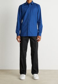 Man med långärmad blå skjorta, svarta byxor och vita sneakers som står mot en enfärgad ljus bakgrund.