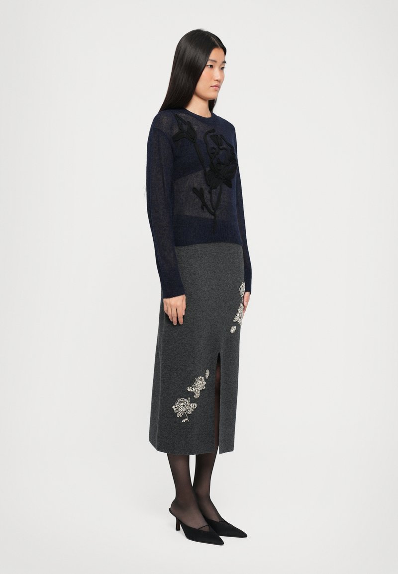 Maglione lavorato a maglia blu navy con pannelli trasparenti e ricami floreali neri, abbinato a una gonna grigia con applicazioni floreali e uno spacco anteriore.