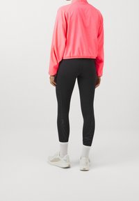 Kirkas vaaleanpunainen urheilu takki pitkillä hihoilla ja joustavalla helmalla, yhdistetty mustiin leggingseihin, joissa on kiiltävät sivupaneelit, sekä valkoisiin lenkkareihin.