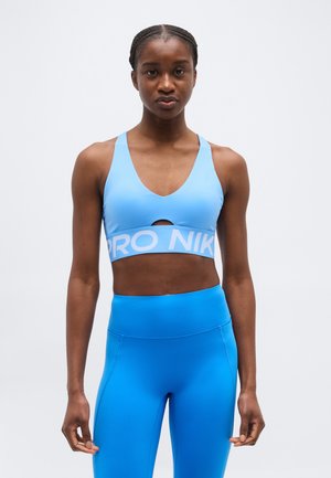 Ung kvinde iført en lyseblå sports-bh og knaldblå højtaljede leggings, stående mod en ensfarvet hvid baggrund.