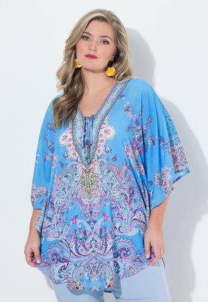 Femme portant une tunique bleue à motifs floraux avec un col lacé et des boucles d'oreilles jaunes en forme de fleur, debout devant un fond blanc.