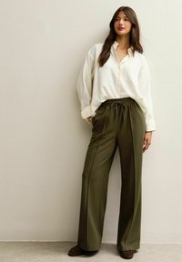 Witte blouse met knoopsluiting, lange mouwen en zijzak. Olijfgroene wijde broek met trekkoord in de taille. Bruine textuur instappers.