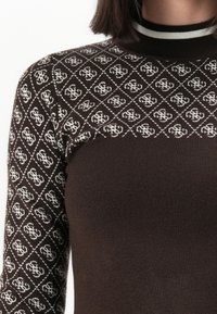 Mujer vistiendo un suéter marrón con patrones geométricos blancos en las mangas y la parte superior del pecho, incluida una detalle de cuello a rayas.