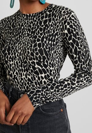 Mustavalkoinen, kirahvin kuvioinen pitkähihainen paita, yhdistetty korkeavyötäröisiin tummiin farkkuihin ja teal-sinisiin roikkuviin korvakoruihin, kädet lepäävät lähellä vyötäröä.