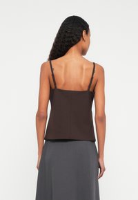 Sieviete ar gariem cirtainiem matiem, tērpta bezgrumbu brūnā blūzē ar plānām siksnām un pelēkā augstās vidukļa svārkos, skatīta no aizmugures.