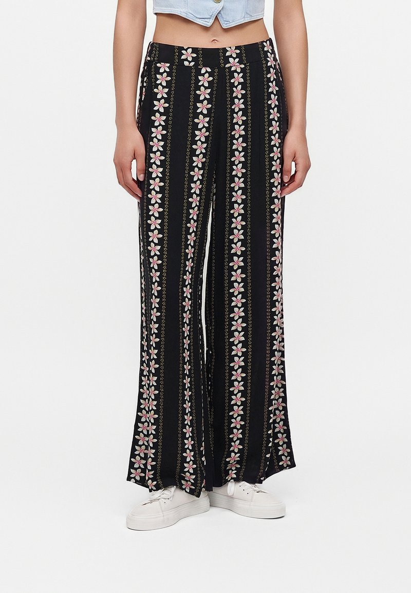 Femme portant un pantalon large noir avec des rayures florales verticales roses et blanches et des baskets blanches, debout devant un fond uni.