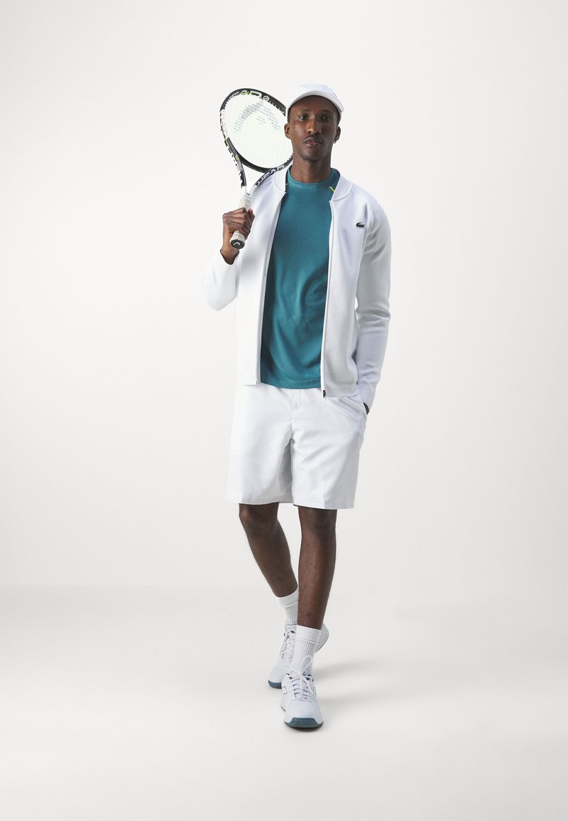 Shorts de sport blancs, t-shirt turquoise, veste zippée blanche et casquette. Tenant une raquette de tennis, présentant des éléments de contraste de texture et de couleur.