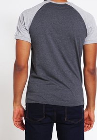 Hombre que lleva una camiseta de manga corta raglán con mangas grises y cuerpo gris oscuro, combinada con jeans oscuros, vista desde atrás.