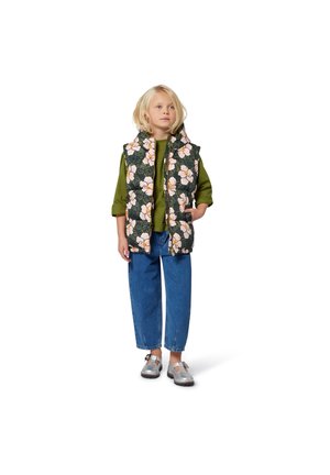 Geverfde bloemenvest in donkergroen met roze bloemen, gelaagd over een groene top, gecombineerd met blauwe baggy jeans en zilveren schoenen.