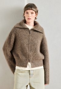 Sweater castanho de mohair com fecho, gola alta, bainha canelada e ombros caídos, sobre uma t-shirt de cor clara. Jeans claros.