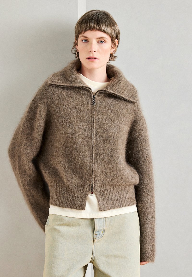 Sweater castanho de mohair com fecho, gola alta, bainha canelada e ombros caídos, sobre uma t-shirt de cor clara. Jeans claros.