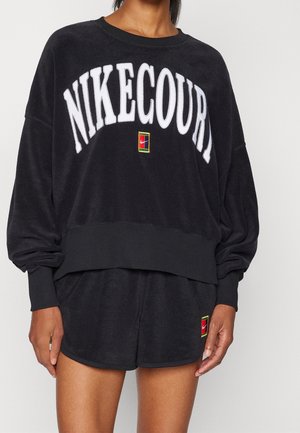 Personne portant un sweat-shirt NikeCourt noir et un short assorti avec un texte de logo blanc et de petits patchs Nike colorés.