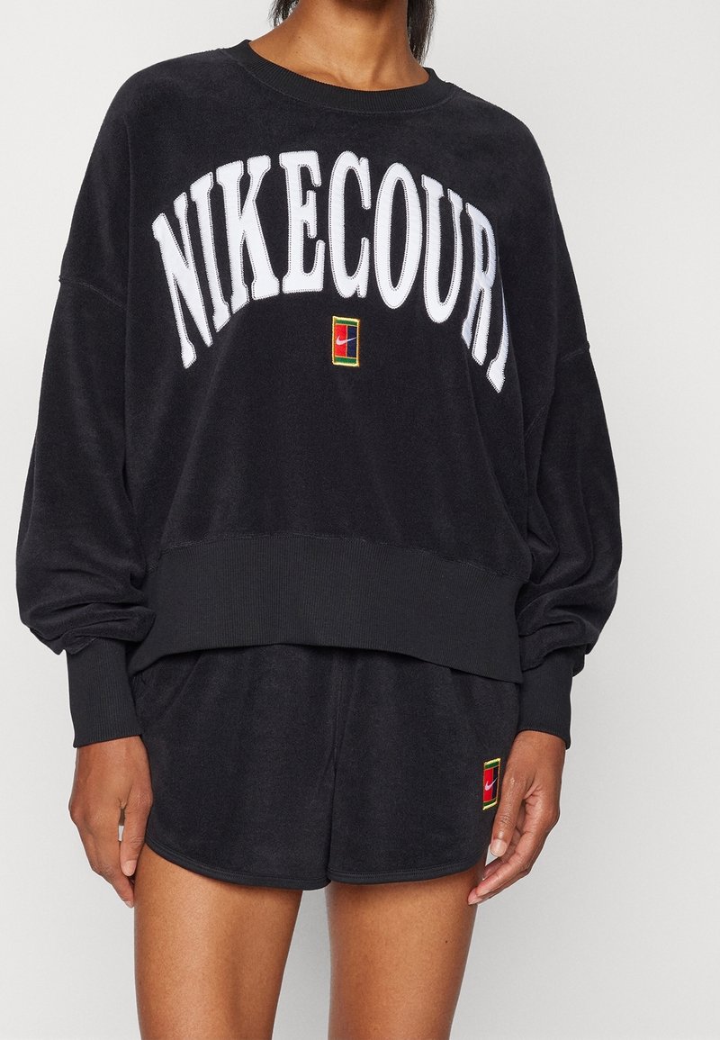 Personne portant un sweat-shirt NikeCourt noir et un short assorti avec un texte de logo blanc et de petits patchs Nike colorés.