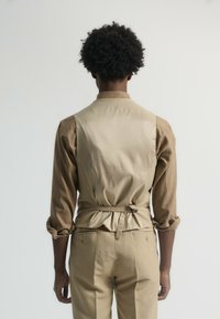 Camisa beige y marrón con un respaldo estructurado, que presenta un detalle de cintura fruncida. Tejido suave con mangas largas remangadas en los puños.