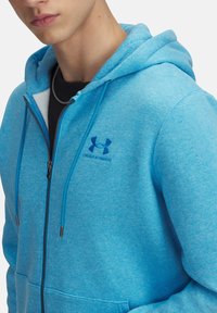 Světle modrá mikina na zip s jemnou texturou, která má přední kapsu, šňůrky na stažení a logo Under Armour na hrudi.