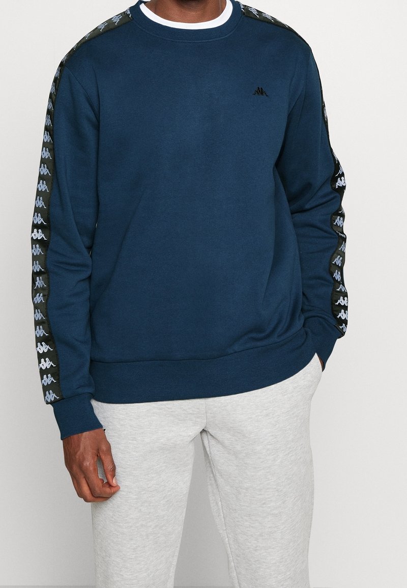 Homme portant un sweat-shirt bleu marine avec un ruban logo blanc sur les manches et un pantalon de survêtement gris clair, debout les mains dans les poches.