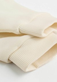 Jasno kremowa dzianina z prążkowanymi mankietami, charakteryzująca się miękką fakturą i złożoną konstrukcją, podkreślająca szczegóły bezszwowej budowy.