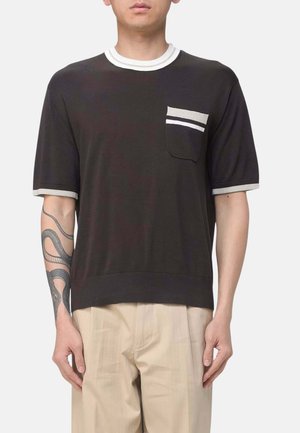 Uomo con camicia nera a maniche corte con tasca e colletto a righe bianche, pantaloni beige e un tatuaggio di serpente sull'avambraccio sinistro.