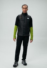 Crni izolirani prsluk preko crne majice dugih rukava s zelenim rukavima, uparen s crnim hlačama i sivim sportskim cipelama.
