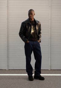 Veste en cuir noire portée sur un t-shirt noir avec un motif doré, associée à un jean bleu foncé et des chaussures noires, sur un fond gris.