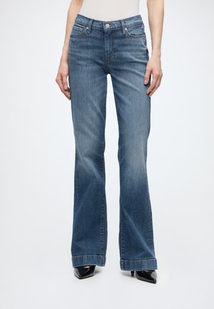 Γυναίκα που φορά μεσαίου χρώματος τζιν bootcut και μαύρες μυτερές γόβες, στέκεται μπροστά σε απλό φωτεινό φόντο.