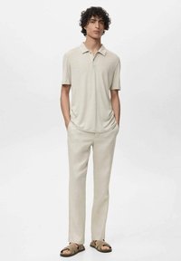 Camisa polo de manga corta beige, combinada con pantalones beige claros y sandalias marrones. Diseño sencillo y corte relajado, con una textura suave.