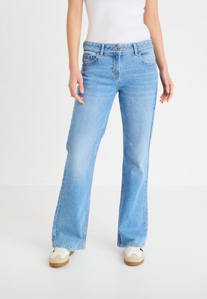 Person trägt hellblaue Jeans mit geradem Bein, weißes Tanktop und weiße Turnschuhe und steht vor einem einfarbigen weißen Hintergrund.