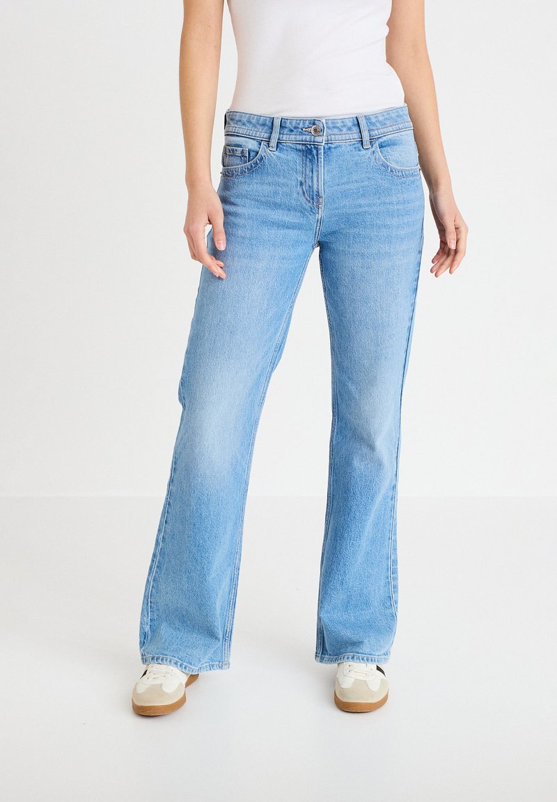 Persona che indossa jeans a gamba dritta azzurri chiari, canottiera bianca e sneakers bianche, in piedi su uno sfondo bianco uniforme.