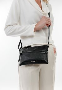 Dámská černá kožená crossbody kabelka s texturovaným povrchem, vybavená zipovým uzávěrem a odnímatelným popruhem, nošená k oblečení v světlých barvách.