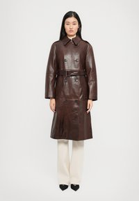 Femme se tient de face, portant un trench-coat en cuir marron foncé ceinturé, un pantalon blanc et des chaussures à talons noirs sur fond uni.