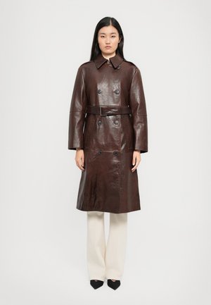 Donna in piedi, rivolta in avanti, indossa un trench coat in pelle marrone scuro con cintura, pantaloni bianchi e scarpe nere a punta, contro uno sfondo neutro.