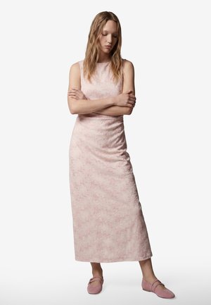 Jeune femme aux longs cheveux portant une robe fleurie rose clair sans manches et des chaussures roses assorties, debout, les bras croisés et regardant vers le bas.