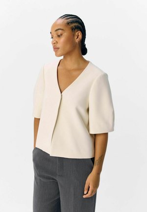 Haut crème à manches courtes avec un décolleté en V, présentant une fermeture à poignées à l'avant et une texture lisse, associé à un pantalon gris ajusté.