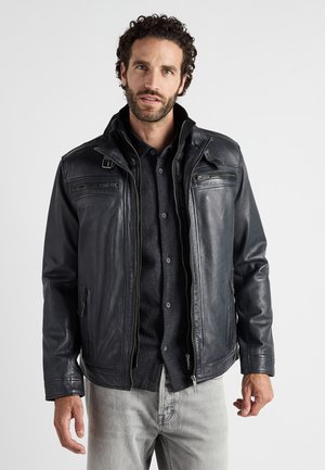 Czarna skórzana kurtka z wysokim kołnierzem, zamkami błyskawicznymi przy kieszeniach, lekko dopasowany krój, noszona na ciemnej koszuli z guzikami, w połączeniu z szarymi dżinsami.