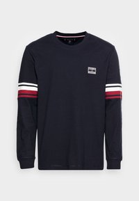 Marineblaues langärmeliges T-Shirt aus Baumwolle, mit weißen und roten gestreiften Akzenten an den Ärmeln und einem kleinen Logo-Patch auf der oberen linken Brust.
