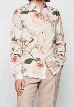 Person wearing a long-sleeve button-up shirt with pink and white rose patterns and beige trousers, one hand in pocket, against a white background.