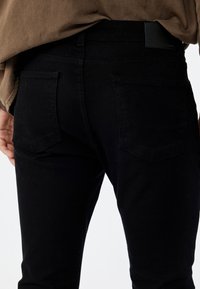 Schwarze Denim-Jeans mit tailliertem Schnitt, ausgestattet mit zwei Gesäßtaschen und einer glatten Textur. Der Bund hat ein Lederpatch in dunkler Farbe.